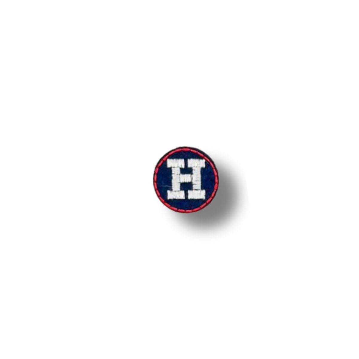 Parche circular con la letra "H" bordada en blanco sobre fondo azul marino, borde rojo fino.